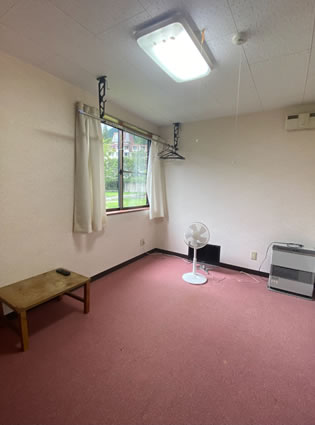 個室寮の部屋、机と扇風機があるシンプルな内装