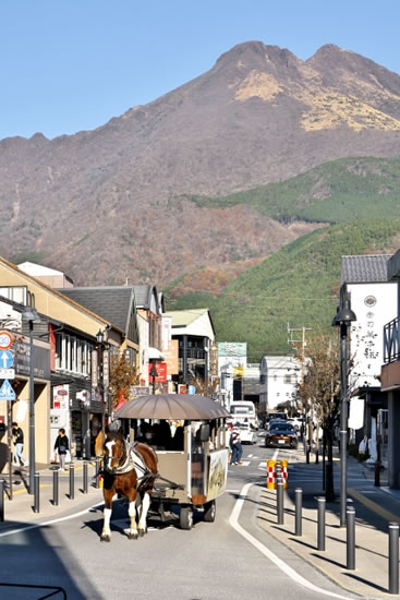 雄大な由布岳を背景に、由布院駅前の通りを乗客を乗せて進む茶色と白の馬が引く観光辻馬車。
